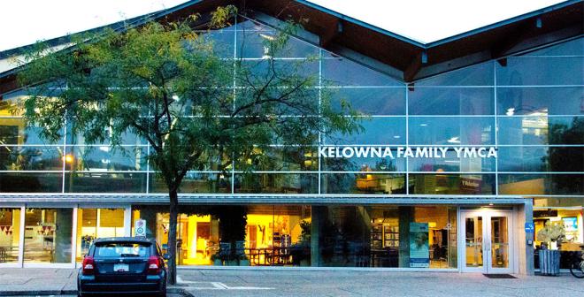 The exterior of the Kelowna Family Y at dawn. The inside is lit up and there are large peaks to the building with windows from floor to ceiling. 