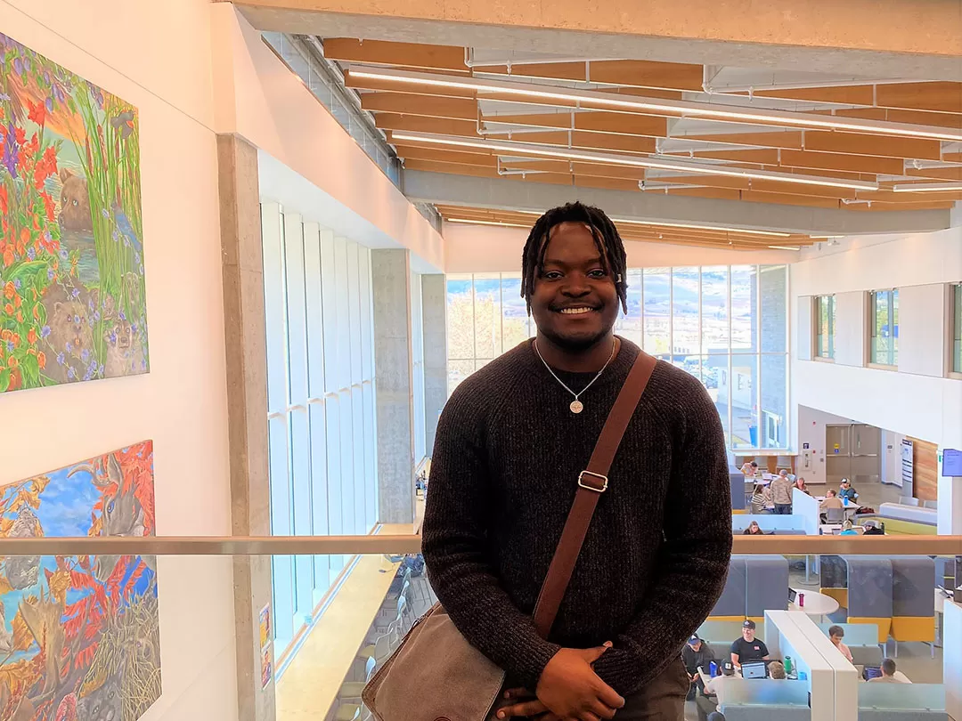 A young man with short black thin dreadlocks in a brown sweater is smiling at the camera in front of a school atrium with paintings, students, and large windows in the background.