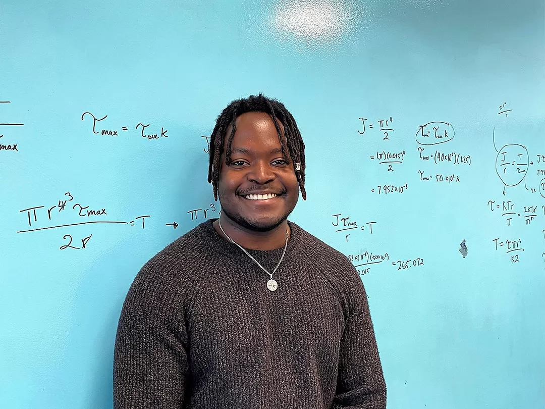 A young man with short black thin dreadlocks in a brown sweater is smiling at the camera in front of a teal board with mathematical equations scribbled on it.