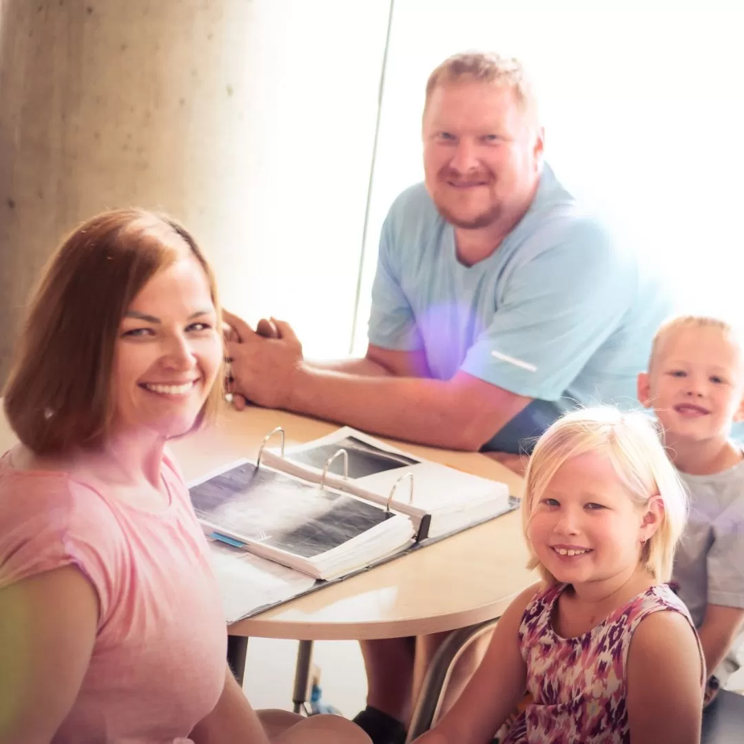 A family of 4 with two young children under the age of 7 are sitting at a table together and smiling at the camera. In front of them is an open binder with photos of x-rays.