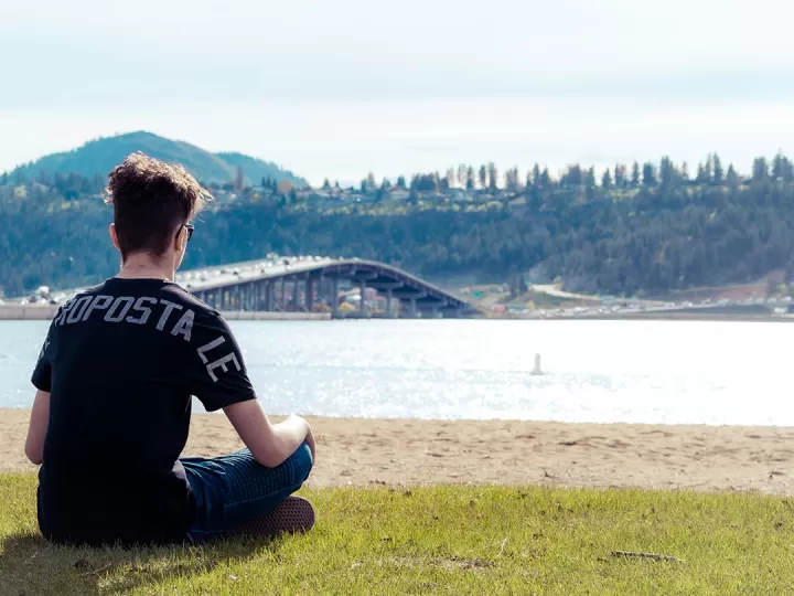 A young man with short dark hair, jeans, sun glasses and a black t shirt is sitting cross legged looking out at a beach. The photo is taken behind him and he is looking out at the water, with a bridge full of cars crossing the lake in the distance. 