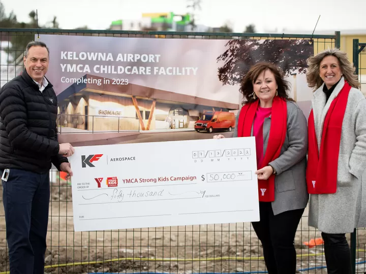 A man in a black jacket, and 2 women in grey winter coats and red YMCA scarves are all standing smiling at a camera holding a large cheque. In the background there is a construction site with a sign on the fence that reads ‘Kelowna Airport YMCA Childcare facility’.